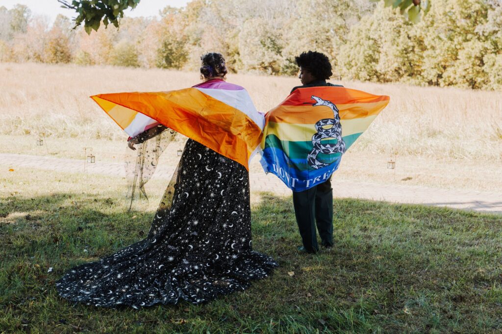 two brides stand in a field wearing pride flags like capes at their Starlight Meadows wedding in North Carolina